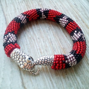 Könnte beinhalten: Ein Armband aus Perlen mit einem Schlangenverschluss. Das Armband ist rot, schwarz und weiß und besteht aus kleinen Perlen. Der Verschluss ist silberfarben und hat eine strukturierte Oberfläche.