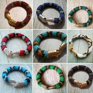 Könnte beinhalten: Neun Perlenarmbänder in Form von Schlangen in verschiedenen Farben und Mustern, jedes mit einem silbernen Schlangenkopfverschluss. Die Armbänder werden auf einer Holzoberfläche präsentiert.