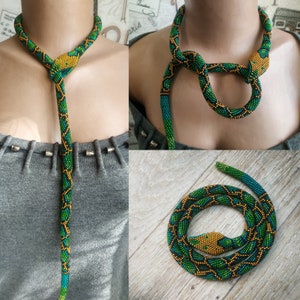 Può includere: Una collana di perline a forma di serpente, con perline verdi, gialle e blu. La testa del serpente è gialla e il corpo è decorato con motivi a rombi verdi e gialli. La collana è mostrata in diverse disposizioni.