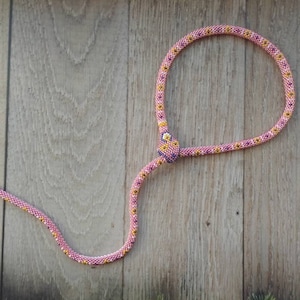 Può includere: Una collana di serpente con perline rosa con una testa di perline gialle e bianche. La collana è su uno sfondo di legno.