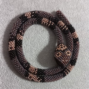 May include: A beaded snake necklace in shades of brown, black, and gold. The snake is coiled, with a detailed head and body. The beads are small and tightly woven, creating a textured surface. The necklace is on a gray background.