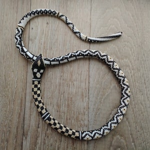 Puede incluir: Un collar de serpiente de cuentas negras y blancas con detalles dorados. La serpiente está enroscada y la cabeza está levantada.