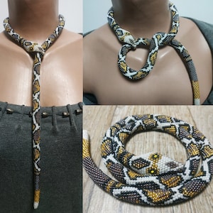 Può includere: Una collana di serpente con perline con un motivo bianco, nero e oro. Il serpente è avvolto attorno al collo e ha una lunga coda che pende sul davanti del corpo.