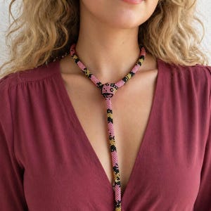 Op de afbeelding: Een kralenketting met een roze, zwarte en gouden patroon. De ketting heeft een Y-vormig ontwerp met een knoop in het midden en een lange, hangende streng. De kralen zijn klein en creëren een textuur.