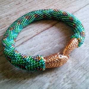 Könnte beinhalten: Ein Perlenarmband in Grün und Gold. Das Armband besteht aus kleinen, runden Perlen in Grün-, Türkis- und Goldtönen. Der Verschluss ist ein goldfarbenes Metall in Form eines Schlangenkopfes.