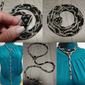 Può includere: Una collana di serpente con perline nere e bianche con un ciondolo a forma di testa di serpente. La collana è mostrata in tre stili diversi: come un choker, come una collana lunga e come un serpente arrotolato.