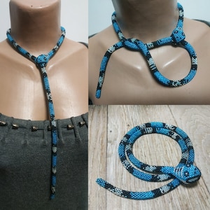 Può includere: Una collana di serpente con perline con un motivo blu e nero. Il serpente è avvolto attorno al collo di una persona e ha una testa con un occhio nero e una lingua bianca.