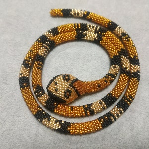 May include: A beaded snake necklace with a black, gold, and white pattern. The snake is coiled and the head is facing the viewer.
