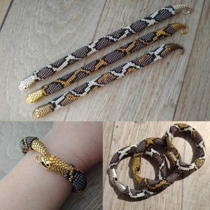 Peut inclure: Trois bracelets en forme de serpent en perles noires, blanches et dorées. Les bracelets sont faits de petites perles et sont conçus pour ressembler à des écailles de serpent. Les bracelets sont présentés sur une surface en bois.
