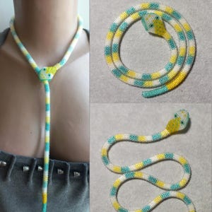 Può includere: Una collana a forma di serpente di perline gialle, bianche e turchesi. La collana è progettata per assomigliare a un serpente, con una testa e un corpo lungo e sottile. La testa del serpente è gialla e il corpo è a strisce con perline bianche, gialle e turchesi. La collana è mostrata in tre diverse viste.