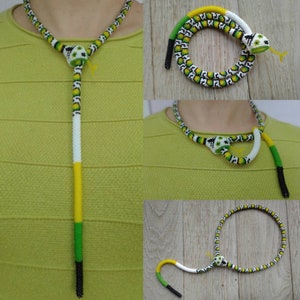 Può includere: Una collana di serpente con perline con un motivo verde, bianco e giallo. Il serpente è avvolto e ha una piccola testa con occhi neri.