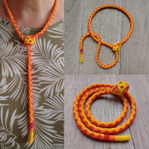 Può includere: Una collana di serpente con perline gialle e arancioni. La collana è realizzata con piccole perline e ha la forma di un serpente. Il serpente è arrotolato e ha una testa con una piccola perlina per il naso.