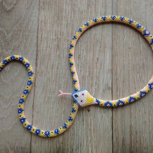 Può includere: Una collana a forma di serpente realizzata con perline gialle, blu e rosa. La testa del serpente è bianca con dettagli blu e la lingua è rosa. La collana è su una superficie di legno.