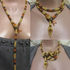 Può includere: Collana a forma di serpente realizzata con perline, mostrata in diverse viste. La collana è principalmente dorata con dettagli neri e arancioni. La testa e la coda del serpente sono decorate con intricate perline. La collana è mostrata come lariat, girocollo e avvolta.
