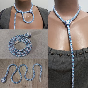 Può includere: Una collana di serpente con perline blu e bianche con un ciondolo a forma di testa di serpente. La collana è mostrata in tre modi diversi: come un choker, come una collana lunga e come un serpente avvolto.