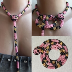 May include: Pink beaded necklace with black and gold detailing. The necklace features a Y-shape and a coiled snake design. The small beads create a textured surface. The necklace is displayed on a mannequin. The necklace's length is not specified.