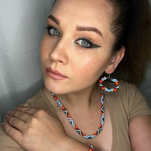 Puede incluir: Un conjunto de collar y pulsera de cuentas con un patrón geométrico en tonos de azul, rojo, blanco y negro. El conjunto incluye un par de pendientes de aro a juego.