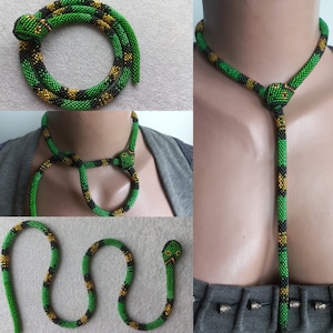 Può includere: Una collana di serpente in perline verdi, nere e dorate. La collana è realizzata con piccole perline ed è a forma di serpente. La testa del serpente è in cima alla collana e la coda in fondo. La collana è mostrata in tre posizioni diverse: arrotolata, indossata intorno al collo e pendente sul davanti del corpo.