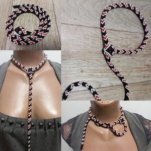 Può includere: Una collana di serpente in perline nere, bianche e rosse. Il serpente è avvolto e ha una testa di perline. La collana è mostrata su un manichino e su una superficie di legno.