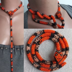 Op de afbeelding: Een kralen slangenketting in oranje, zwart en bruin. De ketting is ontworpen om op een slang te lijken, met een kop en een lange, patroonvormige body. De ketting wordt in meerdere weergaven getoond, waaronder een close-up van de kop en een volledig beeld.