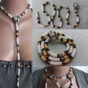 Op de afbeelding: Een kralen slangenketting met een zwart, wit en gouden patroon. De ketting is lang en kan als choker of als langere ketting worden gedragen.