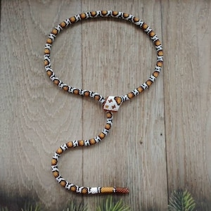 Può includere: Una collana di perline con un design a serpente in marrone, bianco e nero. La collana ha un ciondolo a forma di cubo con un motivo geometrico.