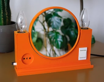 May include: An orange vanity mirror with a round mirror and two light bulbs. The mirror has a white on/off switch and a three-prong outlet. The mirror is on a brown surface.