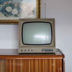 Könnte beinhalten: Ein beigefarbener Retro-Fernseher mit schwarzem Bildschirmrand und ausziehbarer Antenne. Der Fernseher steht auf einem Holzschrank mit glänzender Oberfläche. Der Text "5031" und "lakra" ist auf dem Fernseher zu sehen.