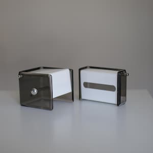 Puede incluir: Dos cajas de pañuelos de acrílico transparente con interior blanco. Los soportes tienen forma rectangular con un panel frontal tintado en gris oscuro y un pomo plateado. Las cajas de pañuelos están sobre una superficie blanca.