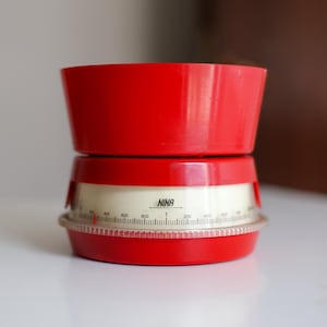 Pode incluir: Uma balança de cozinha vintage vermelha e creme. A balança tem uma tigela vermelha e um mostrador creme com medidas em gramas. A palavra "NINA" está impressa no mostrador. Um acessório de cozinha clássico.