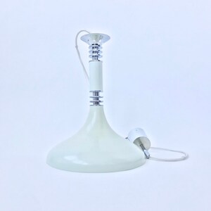 Può includere: Lampada a sospensione color crema con paralume svasato e dettagli cromati. L'apparecchio ha un corpo cilindrico e una parte superiore cromata. Un cavo bianco e un piccolo cilindro bianco sono attaccati alla luce.