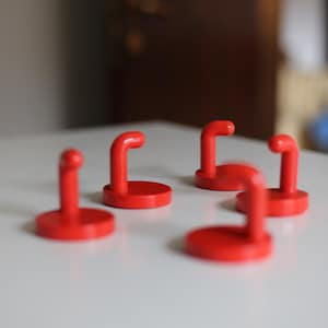 Puede incluir: Cinco pequeños objetos rojos brillantes en forma de gancho con una base circular están dispuestos sobre una superficie blanca. Los ganchos están en posición vertical, con una parte superior curvada. Los objetos están espaciados uniformemente y el fondo está borroso.
