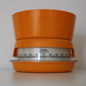 Può includere: Bilancia da cucina vintage arancione con un quadrante di misurazione circolare trasparente. Il quadrante presenta numeri e segni neri, con la parola "POLONA" e una freccia. La bilancia ha un design a più livelli.