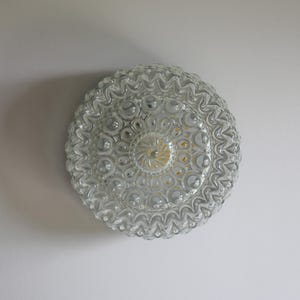 Pode incluir: Uma luminária de teto circular em vidro transparente com uma superfície texturizada, um padrão central de raios de sol rodeado por anéis concêntricos de pontos em relevo e uma borda externa ondulada e festonada que difunde a luz.