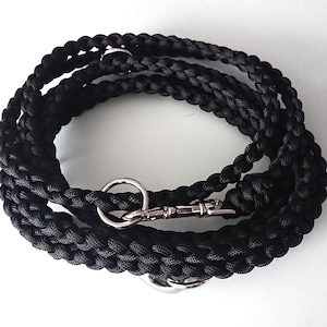 Op de afbeelding: Een zwarte gevlochten hondenriem met zilverkleurige metalen beslag. De riem is opgerold en ligt op een witte ondergrond. Hij heeft een draaibare sluiting en twee metalen ringen.