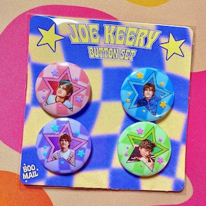 May include: A set of four button badges featuring a person with different coloured backgrounds. The badges are in a star shape and have a white background with a blue, green, pink, and light blue background. The text "JOE KEERY BUTTON SET" is at the top of the card. The text "BOO MAIL" is at the bottom of the card.