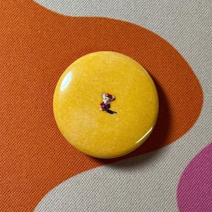 Op de afbeelding: Een ronde gele knop met een kleine cartoonmuis die een rood-wit pakje draagt.
