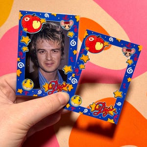 May include: Two blue photo frames with cartoon graphics. One frame holds a photo of a person. The frames have a red apple, yellow stars, a smiling face, and a mouse cartoon. The word "Djos" is written in red and yellow.