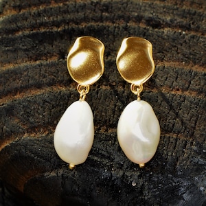 May include: A pair of gold earrings with white pearl drops. The earrings are on a dark brown wooden surface.