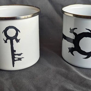 May include: Two white enamel mugs with silver rims and handles. One mug features a black key design, and the other has a black abstract symbol. The mugs are set against a gray background.
