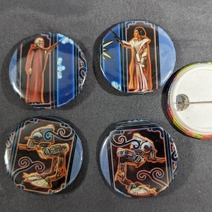 May include: A collection of five round pinback buttons. Four buttons feature illustrations of figures in robes and futuristic vehicles, set against blue and black backgrounds. One button shows the pinback with a rainbow edge.