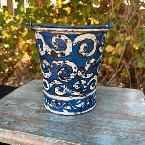 May include: A tapered metal bucket featuring a deep blue base colour with a distressed white painted pattern of repeating swirls and waves. The bucket includes a thin metal handle attached to the top rim, showing a weathered finish throughout.