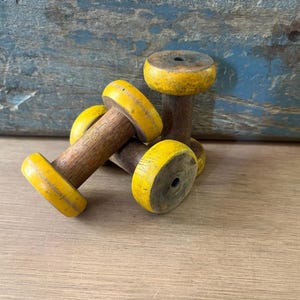 May include: Two wooden spools featuring natural brown cylindrical centres and bright yellow circular ends. Each spool has a small hole centred in the top and bottom discs. The wood grain is visible on the central shafts, while the yellow paint shows signs of wear and texture.