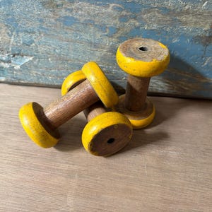 May include: Three vintage wooden spools with natural brown centres and bright yellow painted ends. Each spool features a cylindrical body with a central hole, showing signs of wear and chipped paint on the yellow edges, arranged in a cluster.