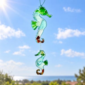 May include: A hanging decoration featuring two seahorses crafted from translucent sea glass in shades of green, blue, and brown. The seahorses are connected by a thin string, with the lower seahorse displaying a small heart-shaped cutout.