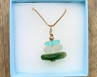 May include: A necklace featuring a gold-tone chain and a pendant composed of three stacked, smooth, rounded pieces of sea glass in shades of light blue, white, and dark green, accented with a small metallic bead at the top of the stack.