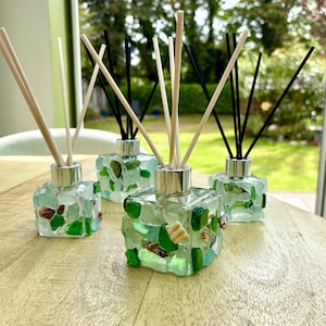 May include: Three square glass reed diffusers decorated with green and clear sea glass pieces and small seashells. Each bottle features a silver-toned collar and contains a mix of light-coloured and black reed sticks extending from the top.