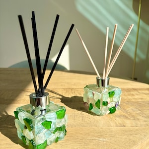 May include: Two glass reed diffusers with black and natural reeds. The square bottles are decorated with green, white, and clear glass pieces. The diffusers sit on a wooden surface. The bottles are approximately 8 cm in height.