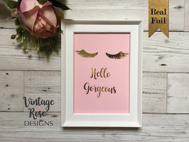 May include: A white framed print with a pink background and gold foil lettering that says "Hello Gorgeous". The print also features two gold foil eyelashes.