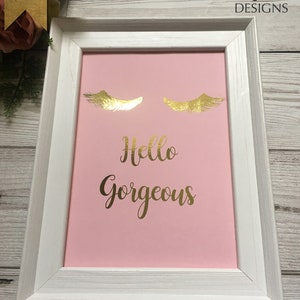 May include: A white framed picture with a pink background. The picture has gold foil eyelashes and the text "Hello Gorgeous" in gold foil.